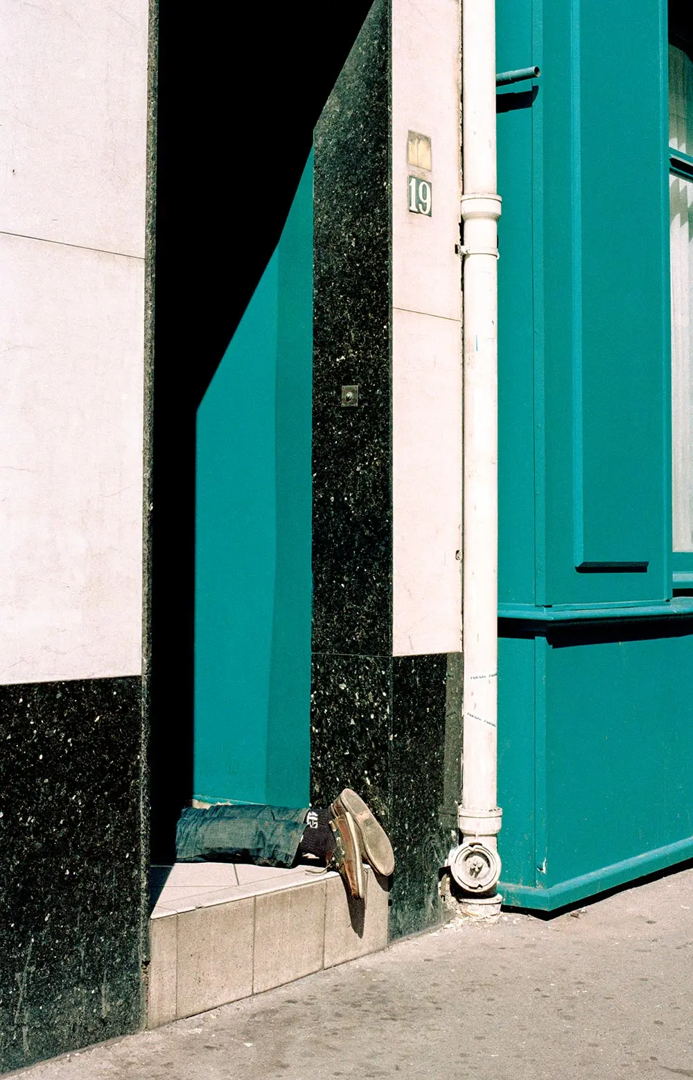 A person lies in a doorway with only their legs and worn shoes visible, surrounded by teal and black walls. Sunlit and marked by the number 19, the scene evokes analog photography, reminiscent of an exhibition "Même pas peur".