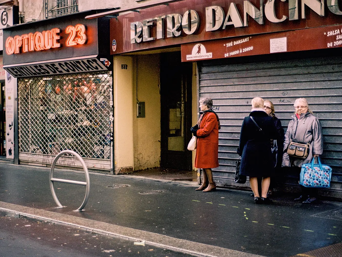 Four elderly women stand on a Paris sidewalk in front of a closed “Retro Dancing” club and next to an optical store with a neon “Optique 23” sign. The wet street adds to the quiet, urban atmosphere.