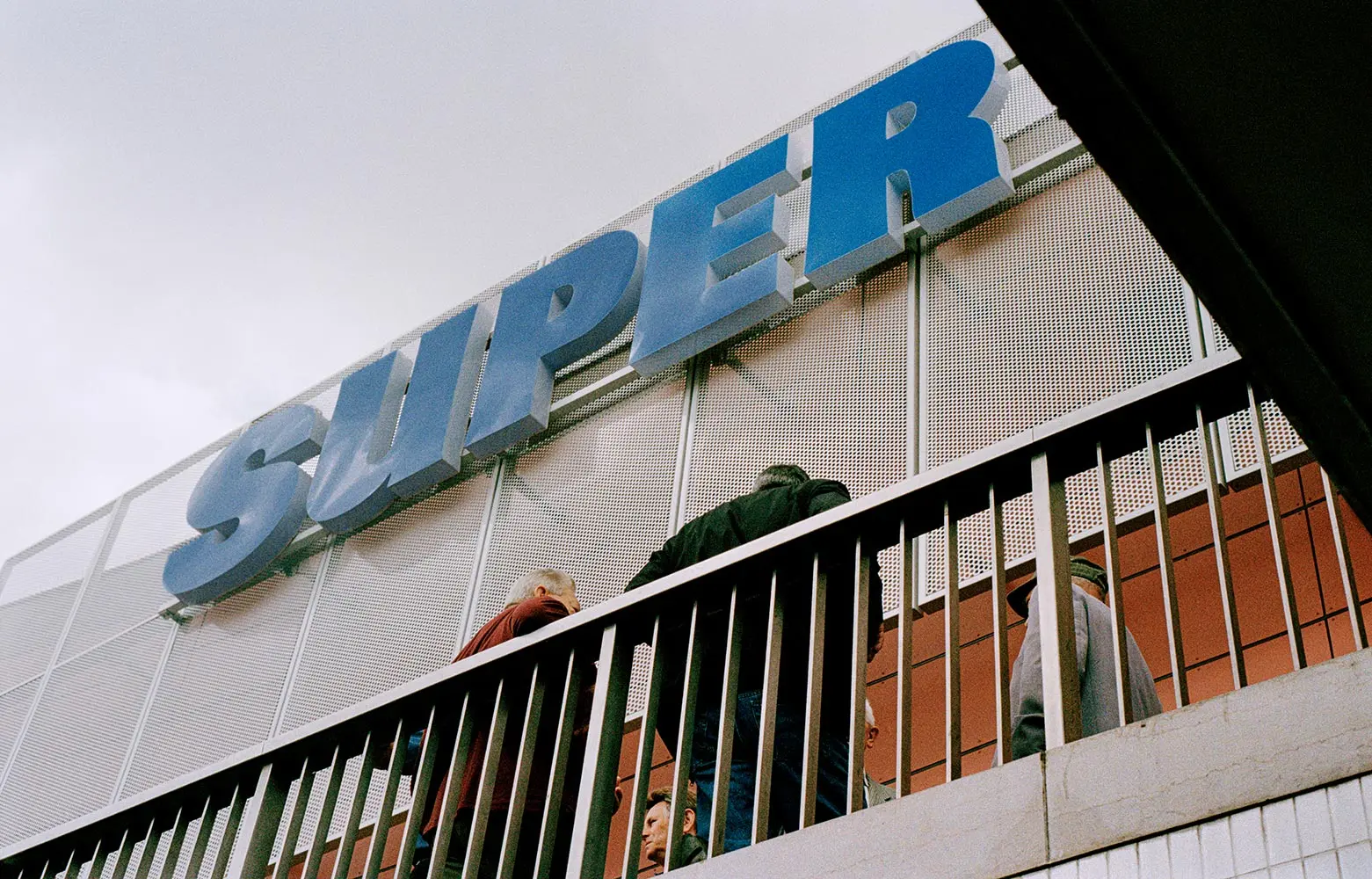 A large blue “SUPER” sign is mounted on a building’s exterior above a metal railing, with several people walking up stairs beneath it, seen from a low angle against a cloudy sky.