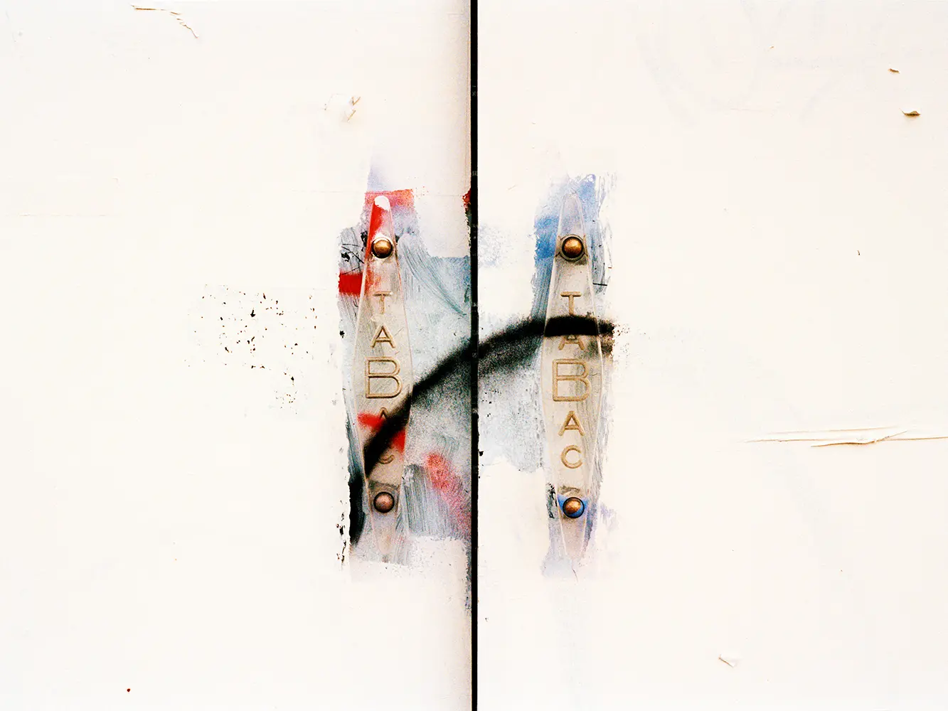 A white wall with two metal plates labeled TABAC, marked by paint smudges and a black spray-painted curved line across them, creates an abstract feel. The otherwise blank wall features only minor scratches and spots.